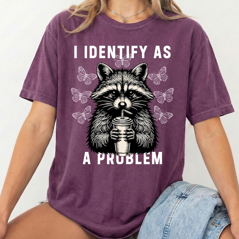 May include: A dark purple short-sleeve t-shirt featuring a black and white graphic of a raccoon holding a drink cup. The text "I IDENTIFY AS A PROBLEM" is printed in white above and below the raccoon, surrounded by several small butterflies.