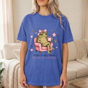May include: A blue t-shirt featuring a graphic of a toad wearing a pink bow, sitting in a pink armchair while holding a pink mug with a heart. The text reads, first of all, Jesus thinks i'm a delight, surrounded by small pink and gold stars.