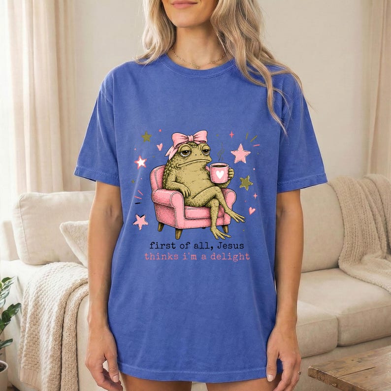 May include: A blue t-shirt featuring a graphic of a toad wearing a pink bow, sitting in a pink armchair while holding a pink mug with a heart. The text reads, first of all, Jesus thinks i'm a delight, surrounded by small pink and gold stars.
