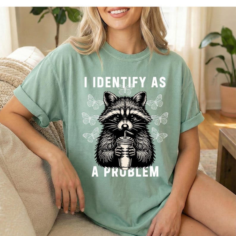 May include: A sage green short-sleeve t-shirt featuring a black and white graphic of a raccoon holding a drink with a straw. The text above the raccoon reads I IDENTIFY AS and the text below reads A PROBLEM, surrounded by small butterfly illustrations.
