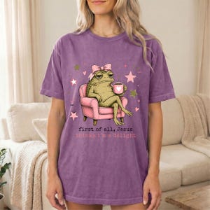 May include: A purple short-sleeve t-shirt featuring a graphic of a green frog wearing a pink bow, sitting in a pink armchair while holding a pink mug with a heart. The text reads, first of all, Jesus thinks i'm a delight, with pink stars.