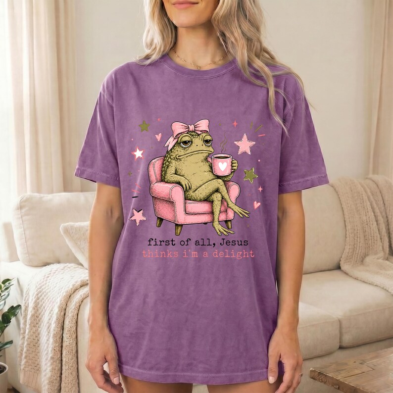 May include: A purple short-sleeve t-shirt featuring a graphic of a green frog wearing a pink bow, sitting in a pink armchair while holding a pink mug with a heart. The text reads, first of all, Jesus thinks i'm a delight, with pink stars.