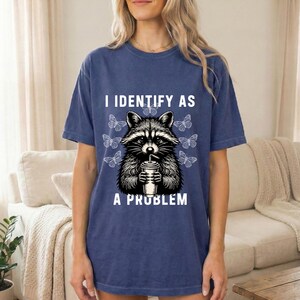 May include: A blue short-sleeve t-shirt featuring a graphic of a raccoon holding a drink with a straw, surrounded by white butterflies. The text I IDENTIFY AS A PROBLEM is printed in white above and below the raccoon illustration.