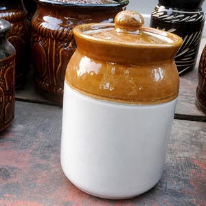 Może przedstawiać: Cylindryczny ceramiczny słoik do przechowywania z błyszczącą białą podstawą oraz kontrastującą górną częścią i pokrywką w kolorze opalenizny. Pokrywka posiada na środku mały, zaokrąglony uchwyt. Słoik ma gładkie, szkliwione wykończenie i szeroki brzeg.