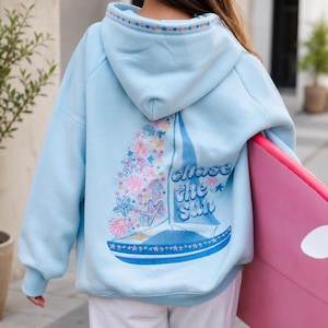 Puede incluir: Sudadera con capucha azul claro con un diseño de velero en la espalda, adornada con conchas marinas y el texto "Chase the Sun". La capucha está adornada con estrellas. La persona sostiene una tabla de surf rosa.