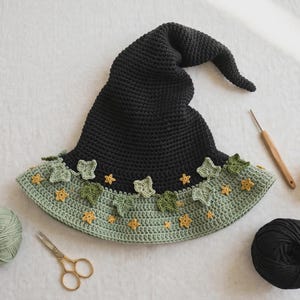 May include: A black crocheted pointed hat with a wide, light green brim decorated with small yellow star-shaped appliques and light green ivy leaf motifs. The hat is surrounded by a ball of sage green yarn, a ball of black yarn, a yellow yarn ball, and scissors.
