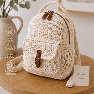 May include: A cream-colored crochet backpack with a front pocket secured by a brown leather strap and buckle. The backpack has a top handle and adjustable straps. The zipper pulls are brown leather. The side of the backpack has a unique floral pattern.