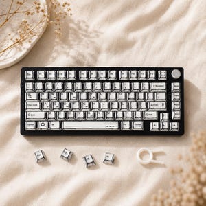 May include: A black and silver mechanical keyboard with white keycaps. The keyboard has a silver rotary knob and several detached keycaps. A white keycap puller is also present. The keyboard is on a beige surface.