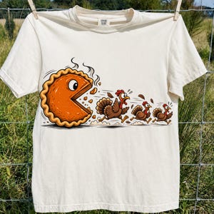 May include: A cream-colored short-sleeve t-shirt featuring a graphic of a large, orange, anthropomorphic pumpkin pie with a wide mouth chasing three brown turkeys. The pie has a single visible eye and is depicted as if it is running.