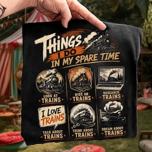 May include: A black t-shirt featuring a graphic with the text "THINGS I DO IN MY SPARE TIME" and six illustrations of trains. The design includes captions: "LOOK AT TRAINS," "RIDE ON TRAINS," "RESEARCH TRAINS," "I LOVE TRAINS," "THINK ABOUT TRAINS," and "DREAM ABOUT TRAINS."