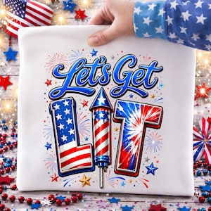 以下が含まれることがあります： Let's Get LITという青い文字がプリントされた白いTシャツ。Lの文字はアメリカ国旗柄で、Iの文字は赤と白のストライプの打ち上げ花火、Tの文字は赤と青の花火の爆発模様がデザインされています。