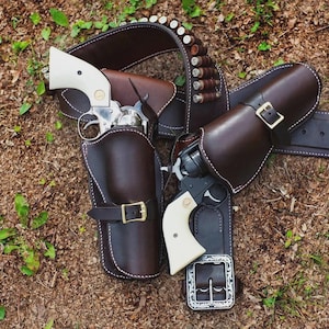 May include: A double leather holster rig featuring two revolvers with cream-colored grips. The dark brown leather holsters have brass buckles and white stitching. A matching leather belt includes loops holding several metallic cartridges.