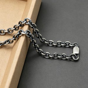 以下が含まれることがあります： 酸化仕上げが施された、暗い色調の円形リンクが連なるシルバーカラーの金属製チェーンネックレス。チェーンの一端には長方形の留め具が備わっており、全体を通して一貫したリンクパターンが特徴的なデザインとなっています。