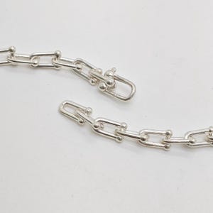 May include: A silver-toned chain necklace featuring elongated rectangular links with rounded corners and small spherical accents at each joint. The chain is shown with a U-shaped clasp mechanism that connects the two ends of the metallic link structure.