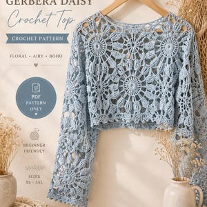 以下が含まれることがあります： 木製のハンガーに掛けられた、水色のGerbera Daisyのかぎ針編みトップス。長袖で、花柄が特徴です。画像には「Crochet Pattern」と「Beginner Friendly」の文字があります。軽やかでボヘミアンな雰囲気です。