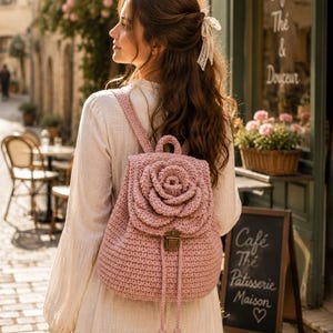 May include: A light pink crochet backpack with a large rose design on the front. The backpack has adjustable straps, a top handle, and a drawstring closure. The bag is made of a soft, textured yarn and has a metal clasp.