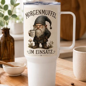 Könnte beinhalten: Ein weißer Edelstahlbecher mit Henkel und durchsichtigem Deckel. Auf der Vorderseite ist ein mürrischer Zwerg mit dunkler Mütze abgebildet, der eine Tasse mit der Aufschrift KAFFEE ERST DANN WELT hält. Darüber steht MORGENMUFFEL und darunter IM EINSATZ.