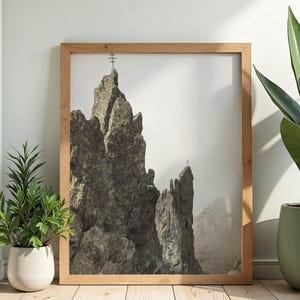 Könnte beinhalten: Ein gerahmter Druck eines zerklüfteten Berggipfels mit einem Kreuz auf dem Gipfel. Das Bild ist in einem hellen Holzrahmen gefasst und wird in Innenräumen ausgestellt. Der Berg ist grau und braun, mit einem dunstigen Himmel im Hintergrund.