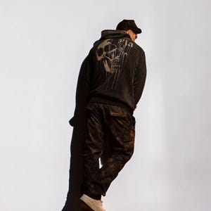 Puede incluir: Una sudadera negra con capucha que presenta un gr&aacute;fico grande y descolorido de una calavera en la espalda con el texto "Pla" encima. La sudadera se combina con pantalones cargo de camuflaje oscuro y una gorra negra, con zapatillas claras.