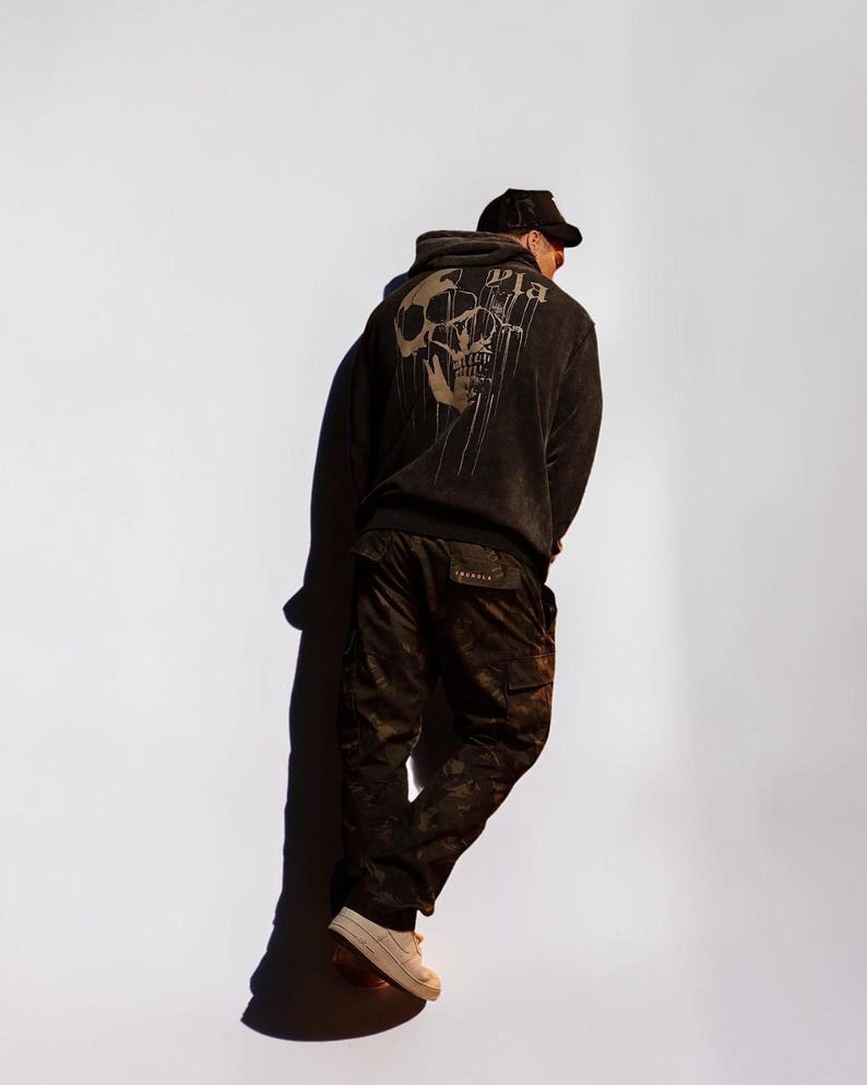 Puede incluir: Una sudadera negra con capucha que presenta un gr&aacute;fico grande y descolorido de una calavera en la espalda con el texto "Pla" encima. La sudadera se combina con pantalones cargo de camuflaje oscuro y una gorra negra, con zapatillas claras.