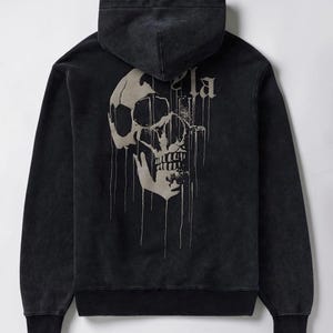 Puede incluir: Una sudadera negra de manga larga con capucha y aspecto desgastado. La parte trasera presenta un gran gr&aacute;fico de calavera beige que gotea, con el texto parcial "la" visible encima. La prenda incluye capucha, pu&ntilde;os acanalados y dobladillo.