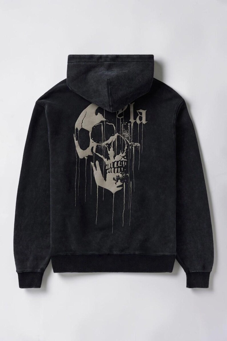 Puede incluir: Una sudadera negra de manga larga con capucha y aspecto desgastado. La parte trasera presenta un gran gr&aacute;fico de calavera beige que gotea, con el texto parcial "la" visible encima. La prenda incluye capucha, pu&ntilde;os acanalados y dobladillo.