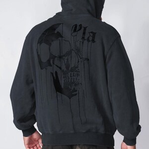 Puede incluir: Una sudadera con capucha negra que presenta un gr&aacute;fico grande de una calavera oscura y goteante impreso en la espalda. El dise&ntilde;o incluye letras estilizadas sobre la calavera. La sudadera tiene mangas largas con pu&ntilde;os y dobladillo acanalados.