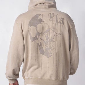 Puede incluir: Una sudadera con capucha de color beige que presenta un gr&aacute;fico grande de una calavera en color beige oscuro en la parte posterior. El dise&ntilde;o de la calavera incluye un efecto de pintura que gotea y el texto "Pla" en una fuente de estilo g&oacute;tico.