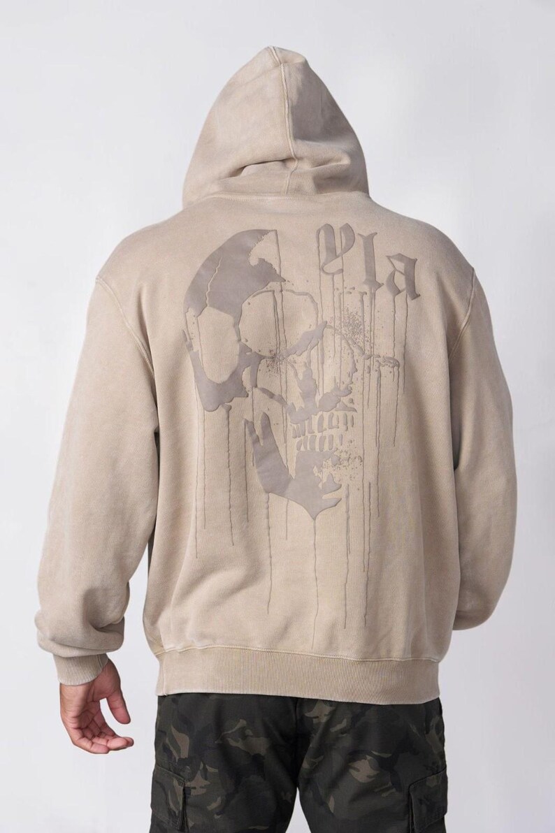Puede incluir: Una sudadera con capucha de color beige que presenta un gr&aacute;fico grande de una calavera en color beige oscuro en la parte posterior. El dise&ntilde;o de la calavera incluye un efecto de pintura que gotea y el texto "Pla" en una fuente de estilo g&oacute;tico.