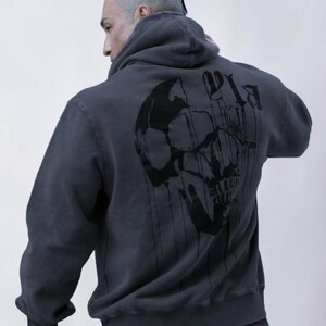 Puede incluir: Una sudadera con capucha de color gris carb&oacute;n que presenta un gran gr&aacute;fico negro de una calavera en la espalda. El dise&ntilde;o incluye l&iacute;neas verticales que parecen gotas que se extienden desde la calavera y letras negras estilizadas.