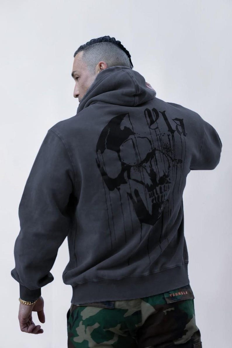 Puede incluir: Una sudadera con capucha de color gris carb&oacute;n que presenta un gran gr&aacute;fico negro de una calavera en la espalda. El dise&ntilde;o incluye l&iacute;neas verticales que parecen gotas que se extienden desde la calavera y letras negras estilizadas.