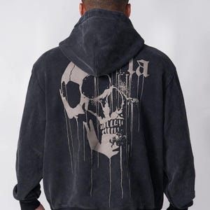 Puede incluir: Una sudadera con capucha de color gris carb&oacute;n que presenta un gr&aacute;fico grande de un cr&aacute;neo de color claro en la espalda. El dise&ntilde;o del cr&aacute;neo incluye l&iacute;neas verticales que parecen gotas, y la tela tiene un aspecto deste&ntilde;ido y desgastado.