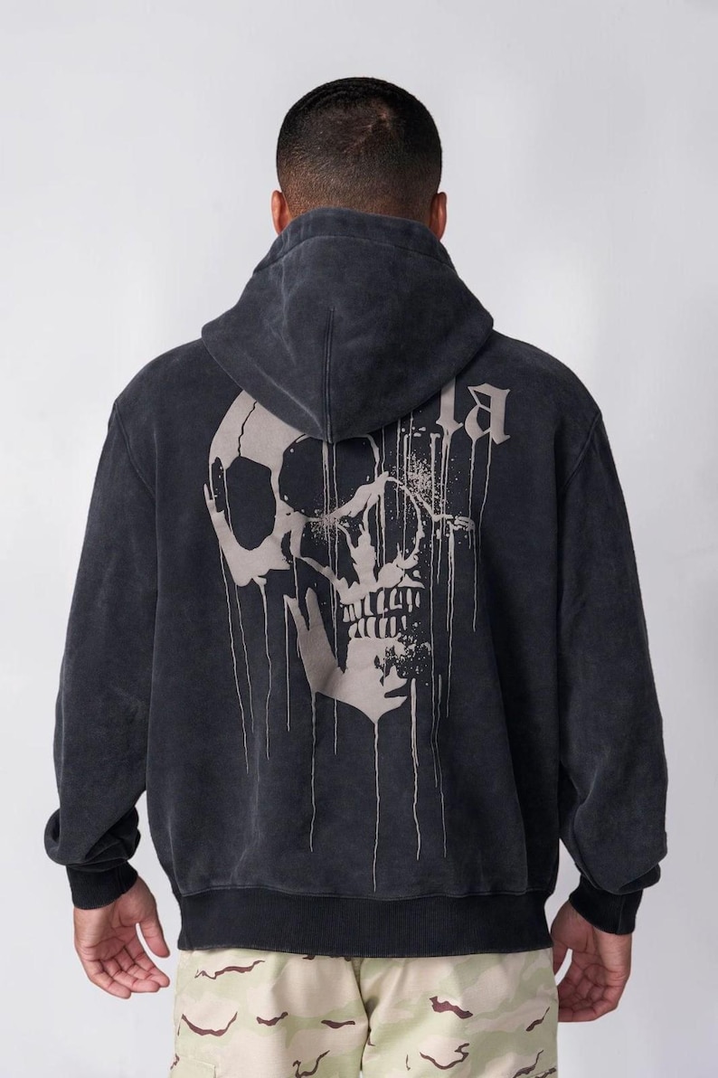 Puede incluir: Una sudadera con capucha de color gris carb&oacute;n que presenta un gr&aacute;fico grande de un cr&aacute;neo de color claro en la espalda. El dise&ntilde;o del cr&aacute;neo incluye l&iacute;neas verticales que parecen gotas, y la tela tiene un aspecto deste&ntilde;ido y desgastado.