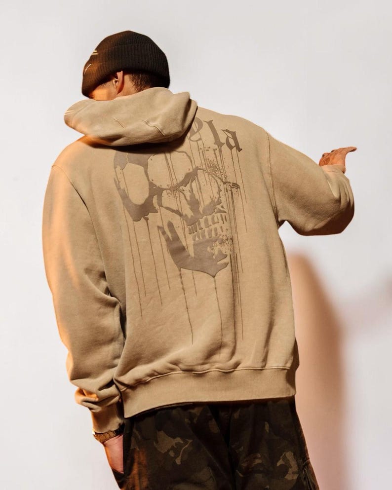 Puede incluir: Una sudadera con capucha de color tostado que presenta un gr&aacute;fico grande de color marr&oacute;n oscuro de una calavera con un efecto de pintura que gotea en la espalda. La sudadera tiene mangas largas, capucha con cord&oacute;n y dobladillo acanalado.