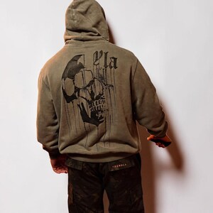 Puede incluir: Una sudadera con capucha de color verde oliva que presenta un gran gr&aacute;fico negro de una calavera con detalles de tinta goteante en la espalda. El texto YLA est&aacute; impreso en una fuente g&oacute;tica sobre la calavera. Se combina con pantalones cargo de camuflaje.