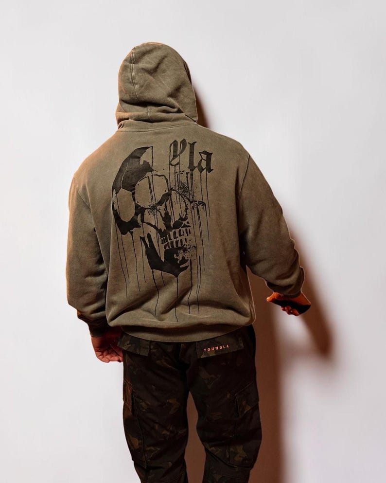 Puede incluir: Una sudadera con capucha de color verde oliva que presenta un gran gr&aacute;fico negro de una calavera con detalles de tinta goteante en la espalda. El texto YLA est&aacute; impreso en una fuente g&oacute;tica sobre la calavera. Se combina con pantalones cargo de camuflaje.