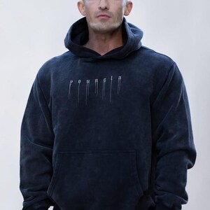 Puede incluir: Una sudadera con capucha y manga larga en color negro carb&oacute;n con una textura deslavada. El frente presenta el texto "youngla" en una fuente de estilo g&oacute;tico con l&iacute;neas verticales que descienden de cada letra y un bolsillo tipo canguro.