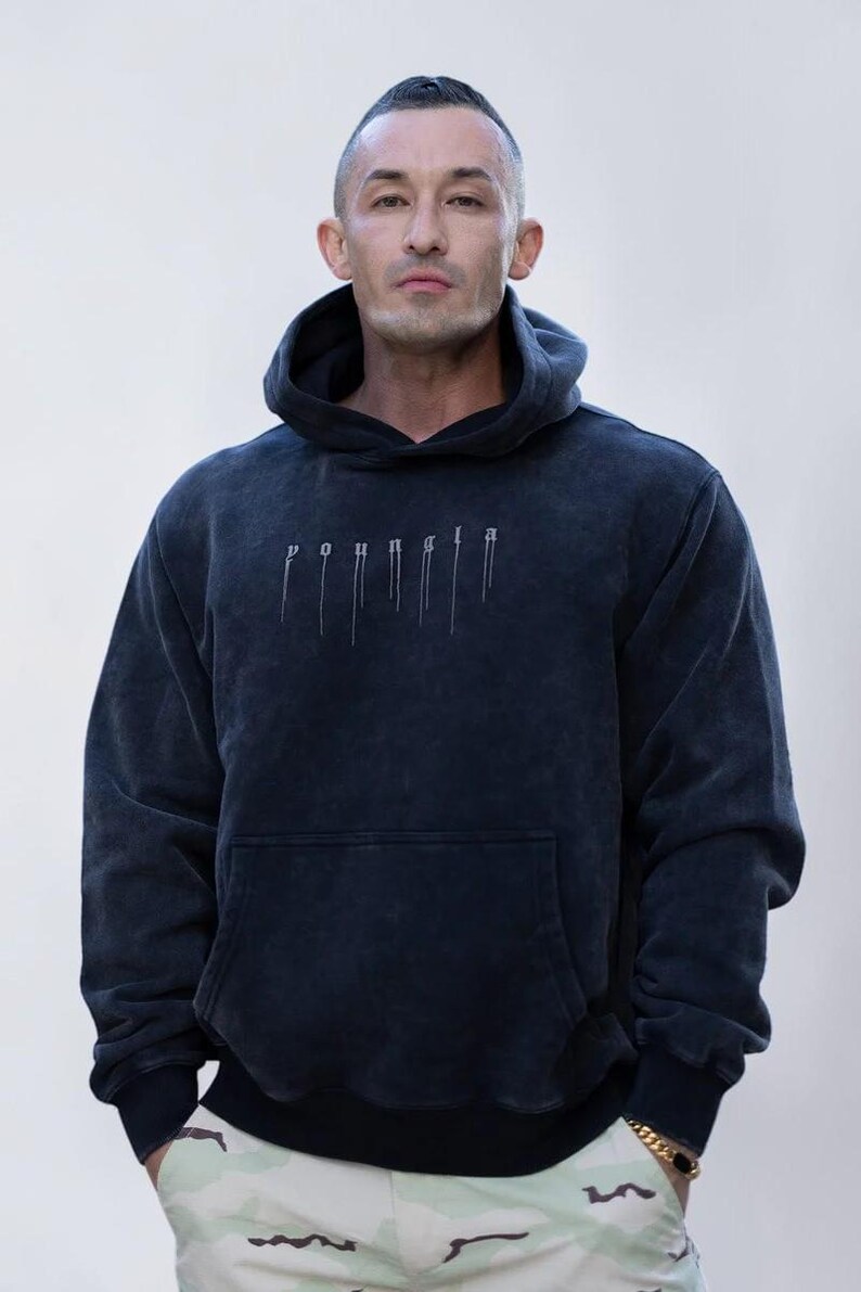 Puede incluir: Una sudadera con capucha y manga larga en color negro carb&oacute;n con una textura deslavada. El frente presenta el texto "youngla" en una fuente de estilo g&oacute;tico con l&iacute;neas verticales que descienden de cada letra y un bolsillo tipo canguro.