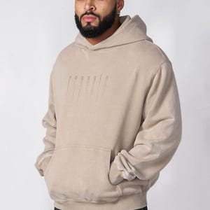 Puede incluir: Una sudadera con capucha de color beige que presenta un bolsillo tipo canguro frontal y un logotipo gr&aacute;fico vertical sutil con efecto de goteo en el pecho. La prenda tiene mangas largas con pu&ntilde;os acanalados y una capucha a juego, combinada con pantalones cargo de camuflaje.