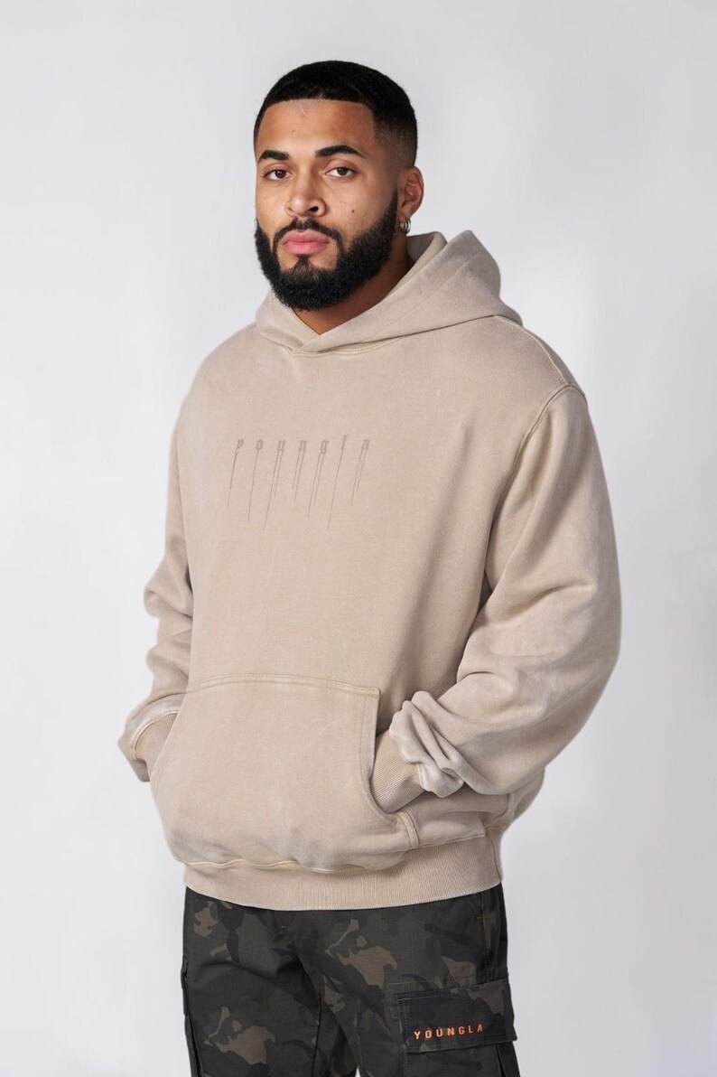 Puede incluir: Una sudadera con capucha de color beige que presenta un bolsillo tipo canguro frontal y un logotipo gr&aacute;fico vertical sutil con efecto de goteo en el pecho. La prenda tiene mangas largas con pu&ntilde;os acanalados y una capucha a juego, combinada con pantalones cargo de camuflaje.