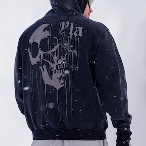 Puede incluir: Una sudadera con capucha negra que presenta un gran gr&aacute;fico gris de una calavera con un efecto de pintura que gotea en la parte posterior. La palabra "Pla" est&aacute; impresa en una fuente g&oacute;tica sobre la calavera. La sudadera se lleva con pantalones gris claro y guantes negros.
