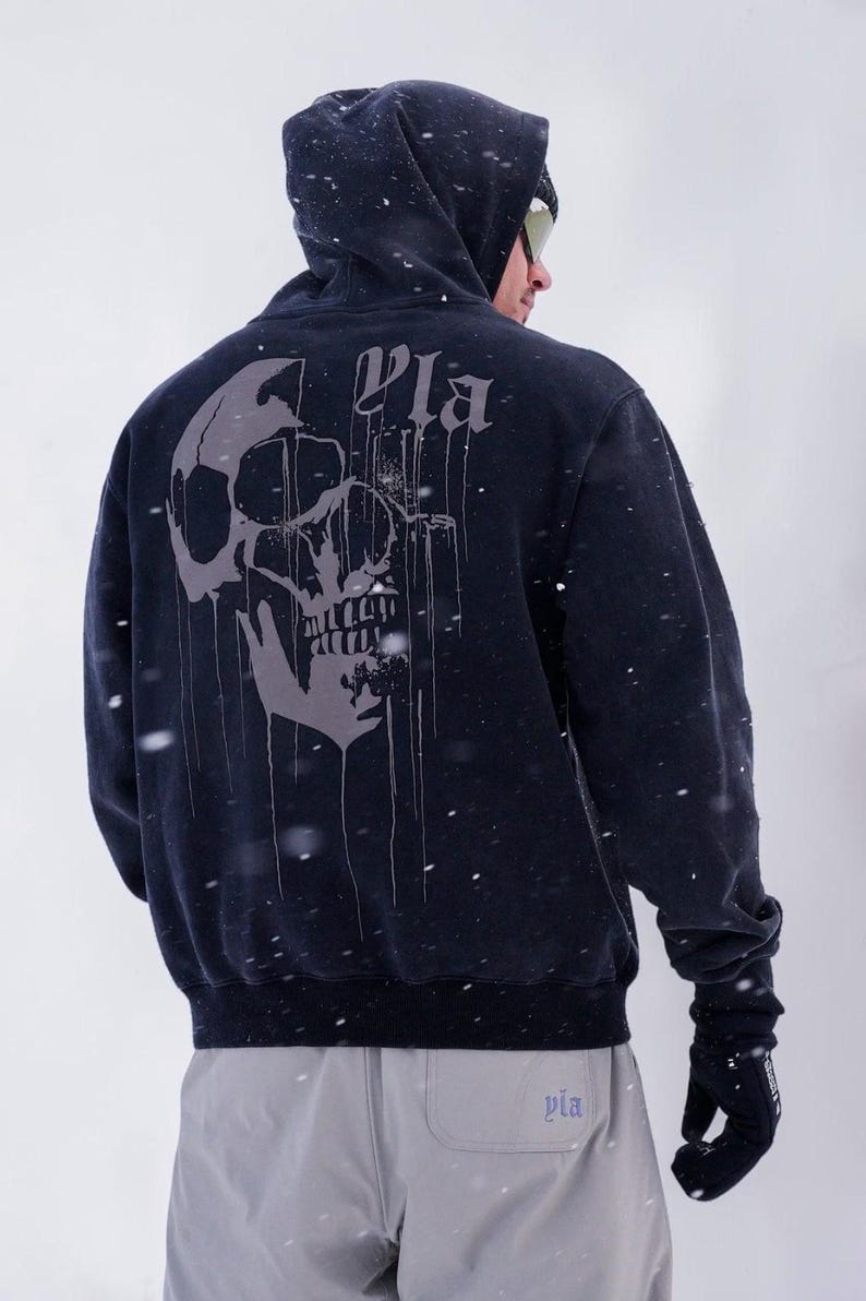 Puede incluir: Una sudadera con capucha negra que presenta un gran gr&aacute;fico gris de una calavera con un efecto de pintura que gotea en la parte posterior. La palabra "Pla" est&aacute; impresa en una fuente g&oacute;tica sobre la calavera. La sudadera se lleva con pantalones gris claro y guantes negros.