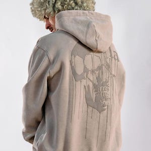 Puede incluir: Una sudadera con capucha de color beige que presenta un gr&aacute;fico grande y oscuro de una calavera en la espalda. El dise&ntilde;o de la calavera incluye l&iacute;neas verticales que parecen gotas que se extienden desde la mand&iacute;bula y las cuencas oculares.