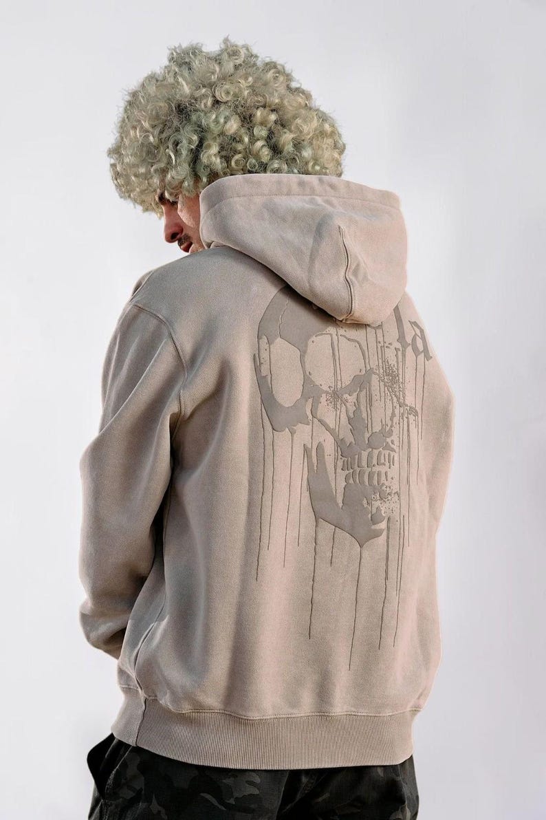 Puede incluir: Una sudadera con capucha de color beige que presenta un gr&aacute;fico grande y oscuro de una calavera en la espalda. El dise&ntilde;o de la calavera incluye l&iacute;neas verticales que parecen gotas que se extienden desde la mand&iacute;bula y las cuencas oculares.