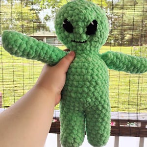 May include: A light green plush alien toy crafted from soft, textured yarn. The figure features a rounded head, two large black oval-shaped eyes, and a small black stitched mouth. It has two outstretched arms and two legs, all made of the same fuzzy material.