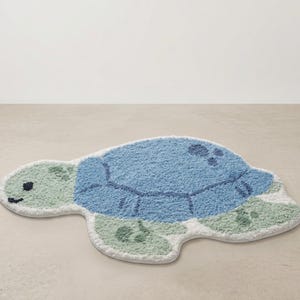 Könnte beinhalten: Eine blau-grüne, schildkrötenförmige Badematte. Die Matte hat ein strukturiertes, weiches Design mit einem hellblauen Panzer, einem grünen Kopf und Flossen sowie einem weißen Rand. Die Schildkröte hat ein lächelndes Gesicht.