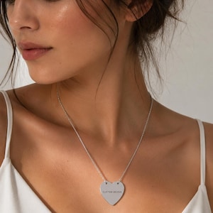 Könnte beinhalten: Halskette mit silbernem herzförmigen Anhänger und der Aufschrift "CUSTOM DESIGN". Die Kette hängt an einer feinen Kette. Das Model trägt ein weißes Oberteil.