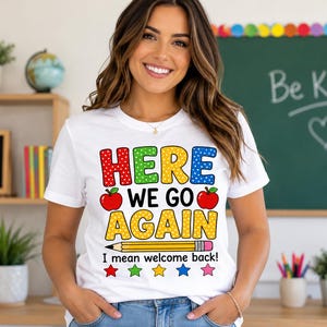 May include: A white short-sleeve t-shirt featuring the text "HERE WE GO AGAIN" in colourful polka-dot letters, two red apples, a yellow pencil, the text "I mean welcome back!" in black, and five small colourful stars at the bottom.