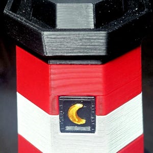 May include: A 3D printed lighthouse model featuring horizontal red and white stripes, a black hexagonal top section, and a light gray cylindrical lantern room. A small black square window displays a gold crescent moon emblem on the front side.