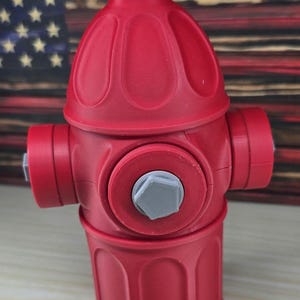 May include: A bright red fire hydrant model featuring a rounded top, three cylindrical side ports, and a central circular face. The hydrant is accented with small, light gray hexagonal caps on the top and the center of the front port.